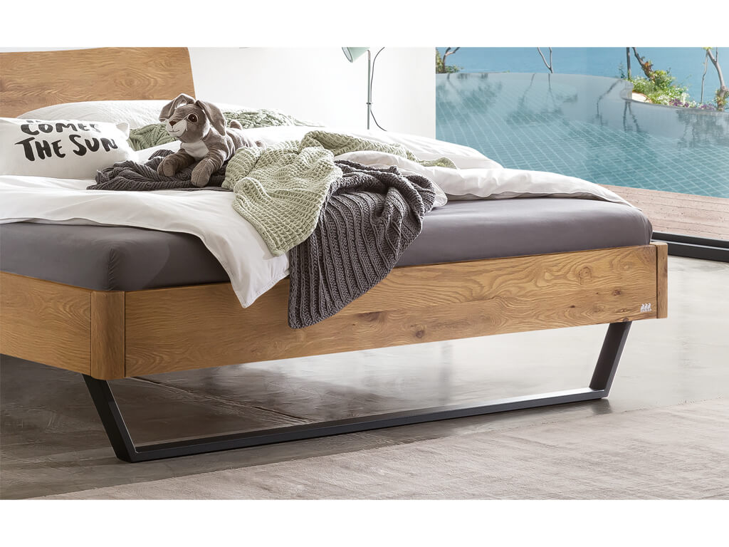 Nahaufnahme der Füße und des Bettrahmens vom Hasena Bett Tresali aus natürlich geölter Wildeiche, das vor einem Fenster mit Pool-Blick steht.