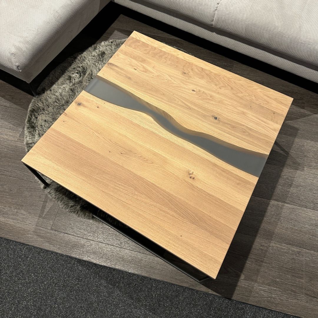 Quadratischer Couchtisch aus Holz mit Glaseinlage, stehend auf einem dunklen Holzboden, umgeben von einem grauen Sofa.