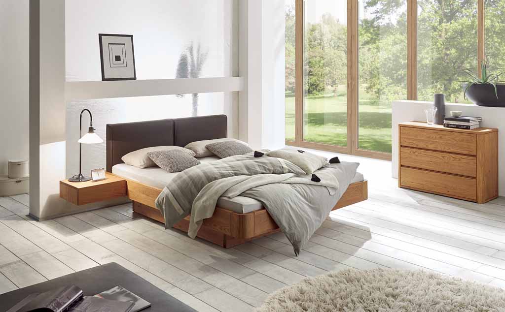 Hasena Oak-Line - Stauraumbett | 180 x 200 cm |  Eiche natur, gebürstet, geölt | Kunstleder dunkelbraun Gemütliches Schlafzimmer mit Holzbett, Nachttisch und Kommode, großer Fensterfront und natürlichem Lichteinfall