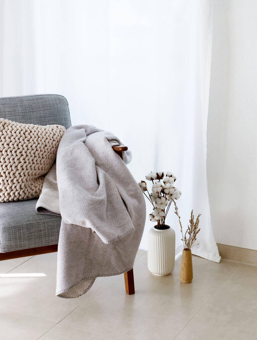 RIEMA. EMMA – Wohndecke, Bio-Baumwolle - meliert | 150 x 200 cm | grau Ein graues Sofa mit Strickkissen und einer hellgrauen Decke. Daneben stehen zwei dekorative Vasen mit Baumwollzweigen und getrockneten Gräsern auf dem Boden.