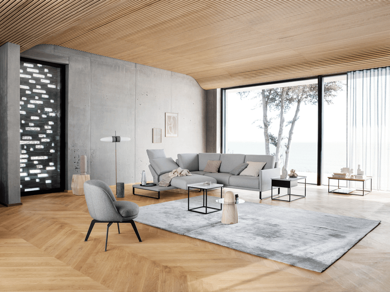 Elegantes Wohnzimmer mit einem großen grauen Sofa, einem Sessel, Holzboden, Glastischen und Panoramafenstern mit Blick auf das Meer.