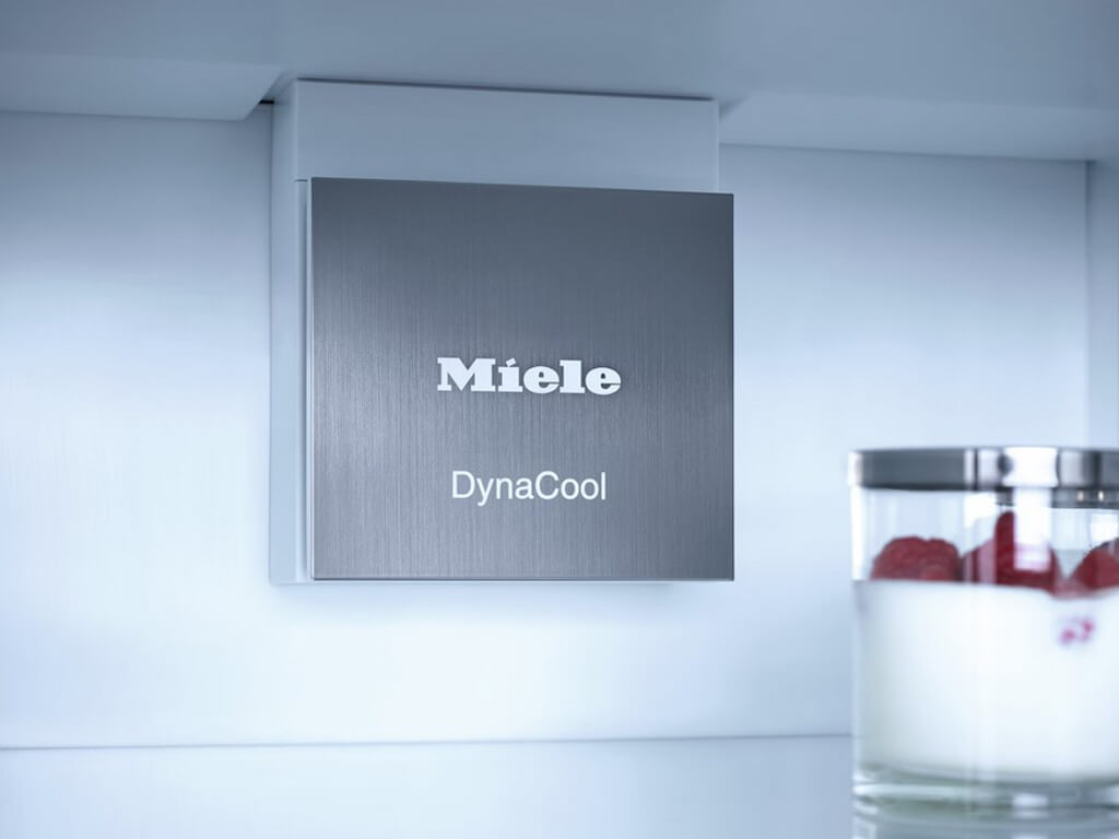 Das Bild zeigt das Innere eines Miele Kühlschranks. Man sieht eine silberne Blende mit den Logos "Miele" und "DynaCool", die sich oben an der Rückwand des Kühlschranks befindet.