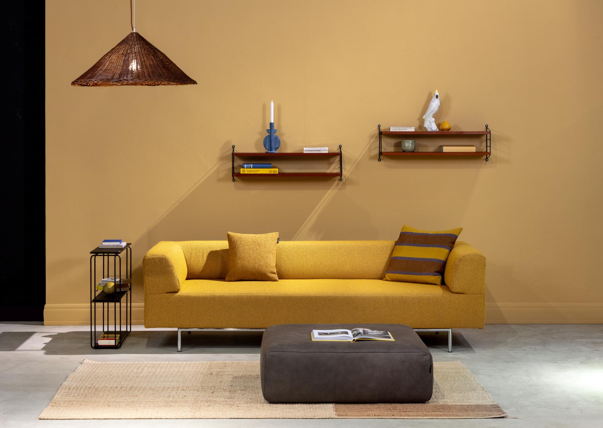 gelbes designersofa mit chromgestell und davor ein brauner polsterhocker stilvoll dekoriert mit kissen und wandregalen in einem modernen helle raum
