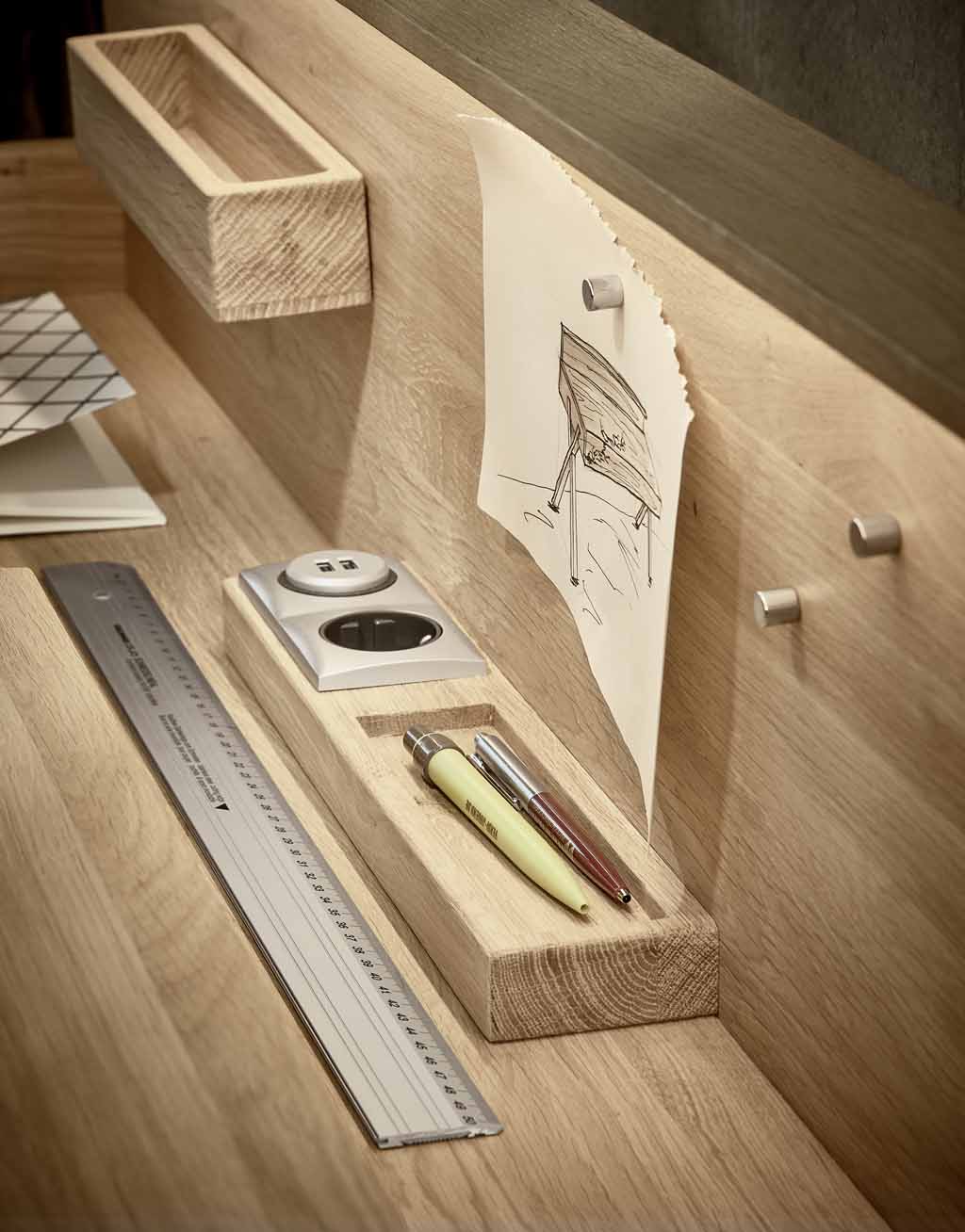Ein Holzschreibtisch mit Zeichenmaterialien, einem Lineal und integrierter Steckdose, neben einer Skizze an der Wand.