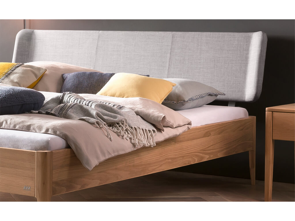 Eine Nahaufnahme des oberen Teils eines Bettes Pienza aus Eiche-bright. Das Bett hat ein gepolstertes Kopfteil in hellem 'Pepe-grey' Stoff und ist mit verschiedenen Kissen in Beige, Grau und Gelb sowie einer gestrickten Decke dekoriert.