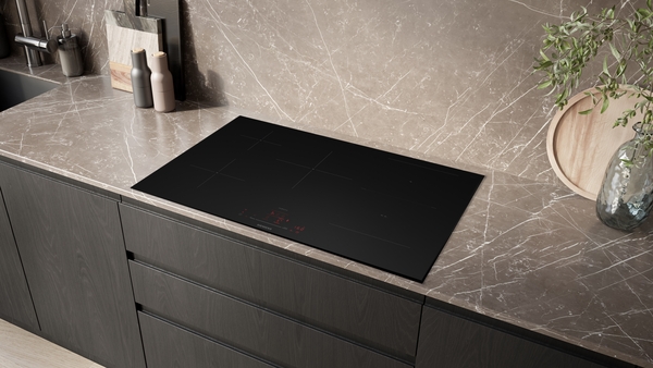 Gesamtansicht des schwarzen Siemens StudioLine Induktionskochfelds (ED87BHWC1E), das flächenbündig in eine braun-marmorierte Küchenarbeitsplatte eingelassen ist. Die Szene zeigt eine moderne Küche mit dunklen Unterschränken.