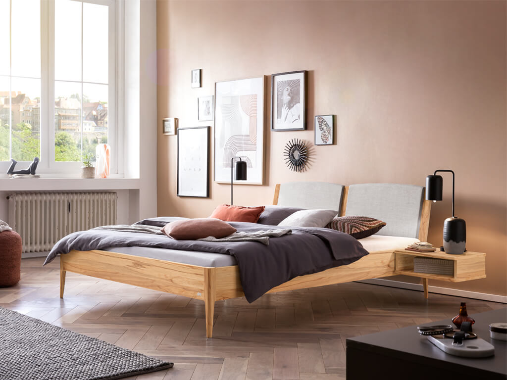Ein helles Schlafzimmer mit einem Hasena Pistoia Bett aus Wildbuche und einem hellgrau gepolsterten Kopfteil. Das Bett ist mit einer dunklen Bettdecke und verschiedenen Kissen dekoriert, die harmonisch zu den Bildern an der Wand passen.