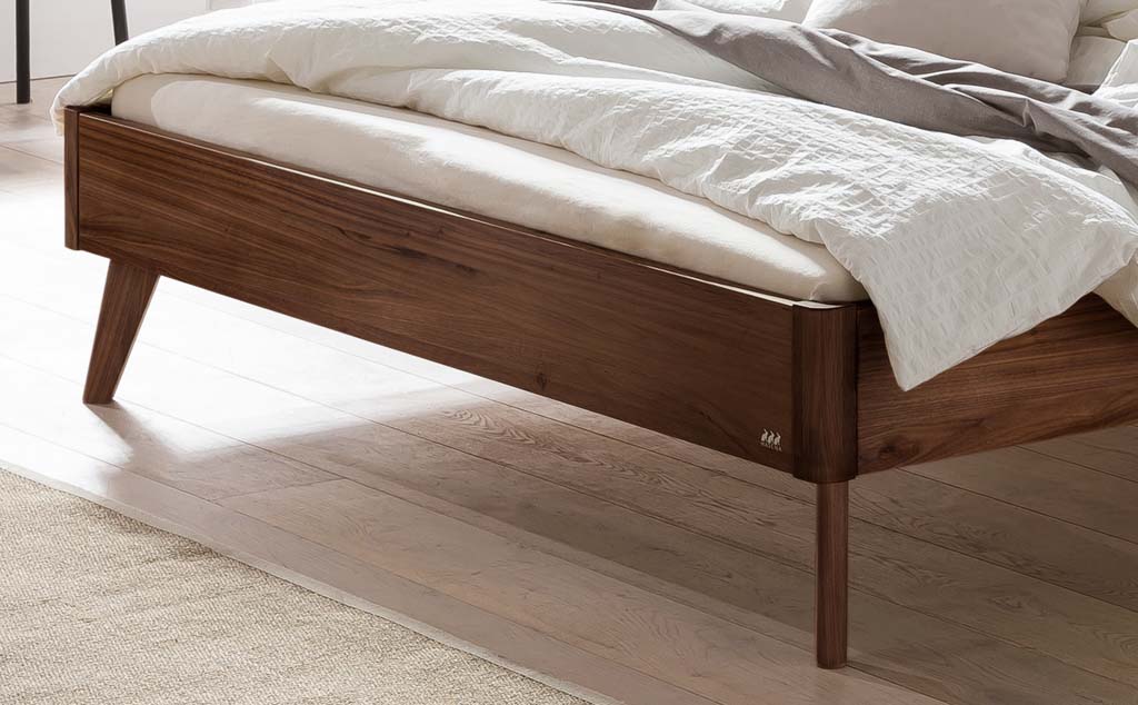 Detailaufnahme eines dunkelbraunen Holzbettes mit Holzfüßen in einem hellen Raum