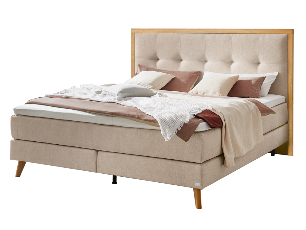 Ein Boxspringbett in hellem Beige mit einem Holzrahmen um das gepolsterte Kopfteil, freigestellt auf weißem Hintergrund.