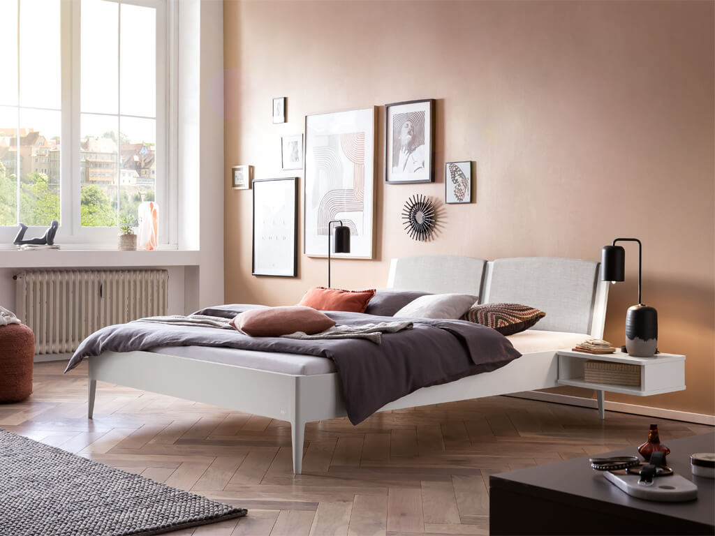Ein stilvolles Schlafzimmer mit einem Hasena Pistoia Bett in weiß lackiertem Holz mit einem gepolsterten Kopfteil aus hellgrauem Stoff. Das Bett ist mit einer dunklen Bettdecke und farbigen Kissen dekoriert, daneben steht ein passender Nachttisch.