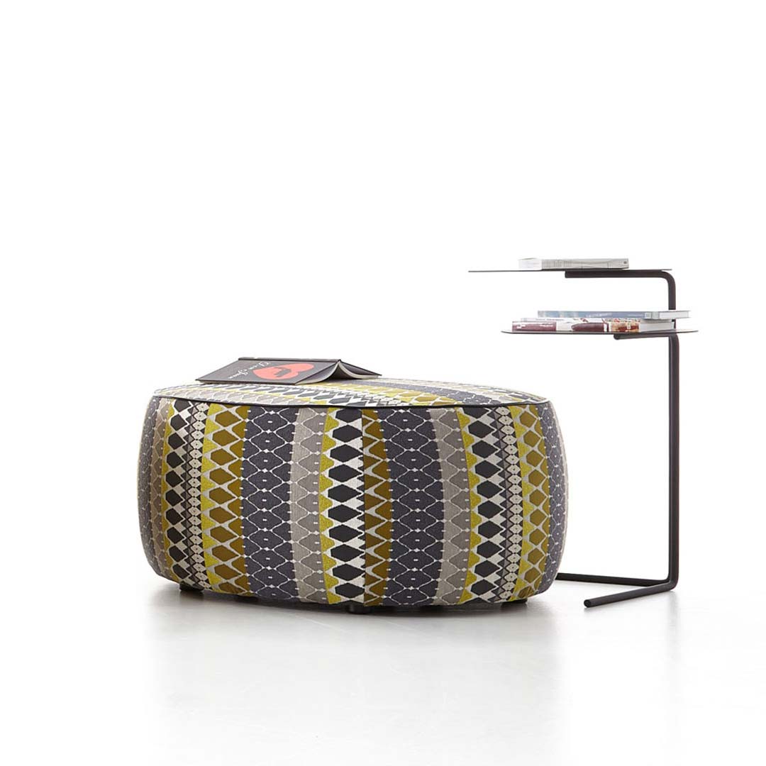 Ein stilvoller Sitzpouf mit geometrischem Muster in Grau, Gelb und Weiß neben einem modernen schwarzen Beistelltisch mit zwei Ablagen.