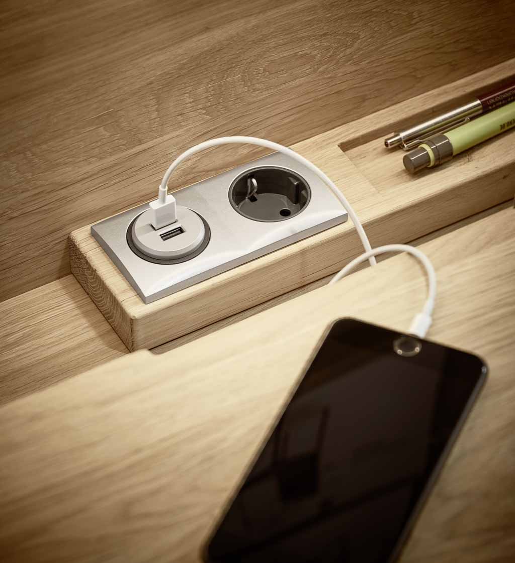 Nahaufnahme einer USB-Steckdosenleiste in einem Holzschreibtisch, an die ein Smartphone angeschlossen ist
