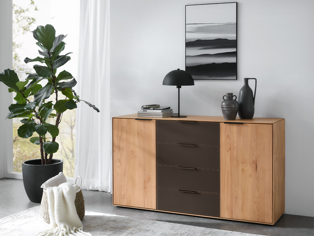 Wöstmann WSM 4400 - Kommode (2150), 4 Schubkästen, 2 Türen | Erle massiv | B: 150,9 cm | konfigurierbar Eine moderne Holzkommode mit schwarzen Schubladen, dekoriert mit Büchern, einer Lampe und Vasen, neben einer großen Zimmerpflanze in einem hellen Raum.