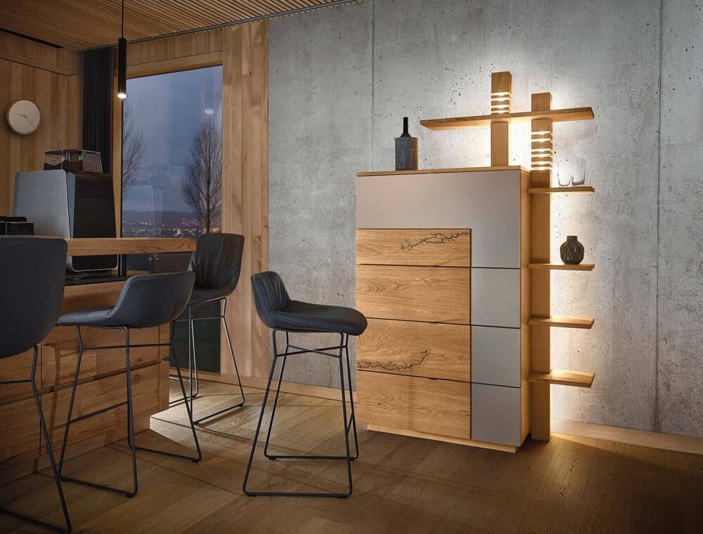 Wimmer Signatura | Highboard | 153125/153126 Innenansicht eines modernen Wohnzimmers mit einem beleuchteten Sideboard aus Holz und Metall, ergänzt durch elegante Barhocker an einem Holztisch.