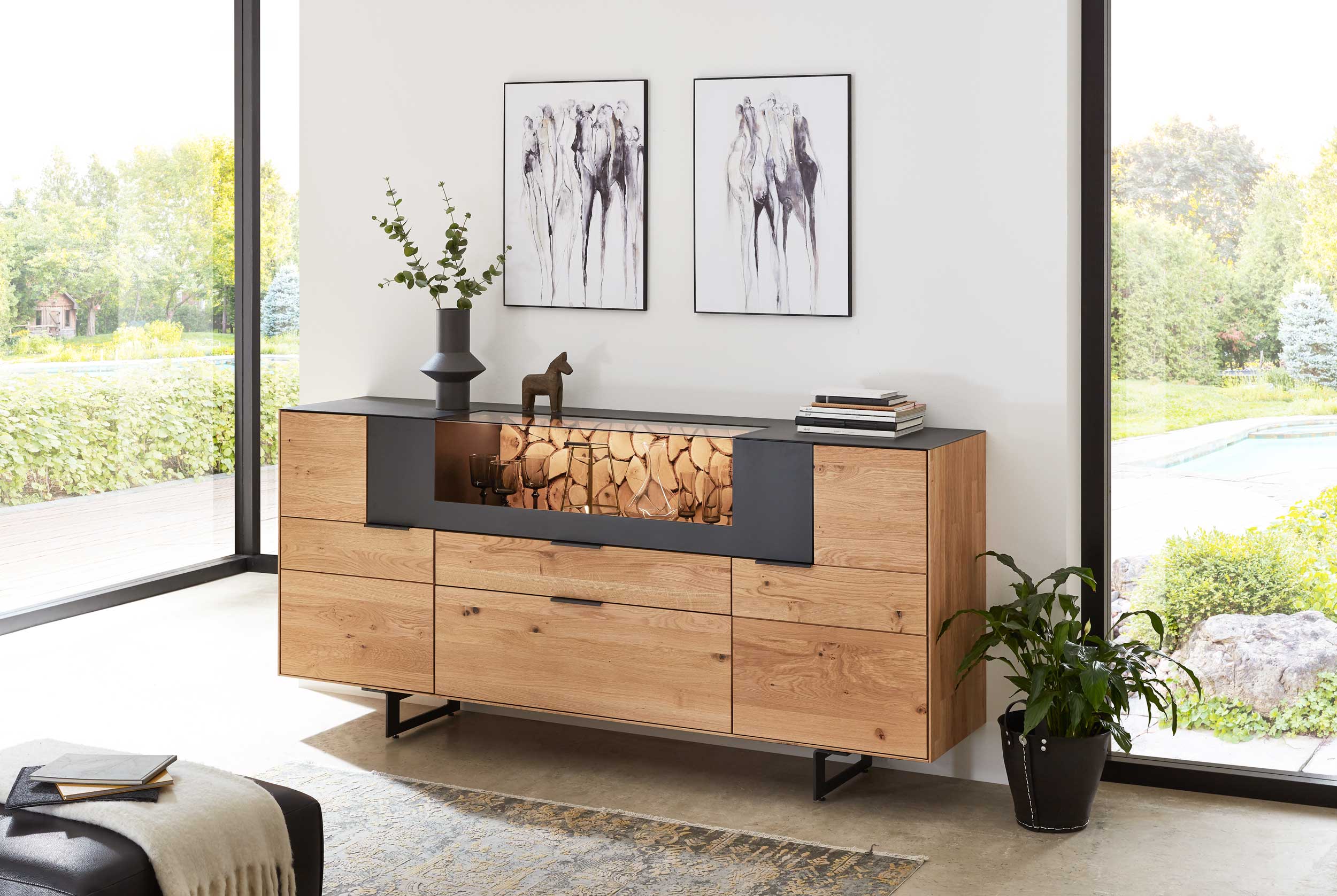 Wöstmann WM 2140 - Sideboard | Type 2040 | B: 209,9 cm | Wildeiche massiv | konfigurierbar Ein moderner Eichenschrank steht in einem hellen Wohnzimmer. Er ist dekoriert mit einer Vase, einen Holztier und Kunstwerken an der Wand. Große Fenster bieten Blick auf einen Garten.