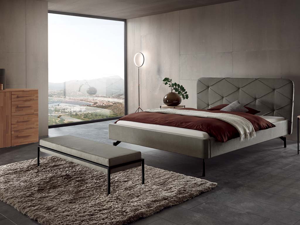 graue Polsterbank auf schwarzem Metallgestell vor einem grauen Polsterbett und auf einem Hochflorteppich in einem modernen SChlafzimmer