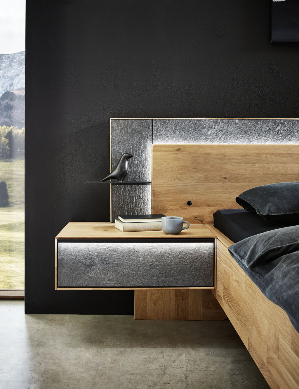 Detailaufnahme eines Bettes mit beleuchtetem Holzkopfteil und schwarzer Metallablage einer beleuchteten Hängekonsole mit grauer Front sowie einer grauer Kopfteilumrandung stilvoll dekoriert