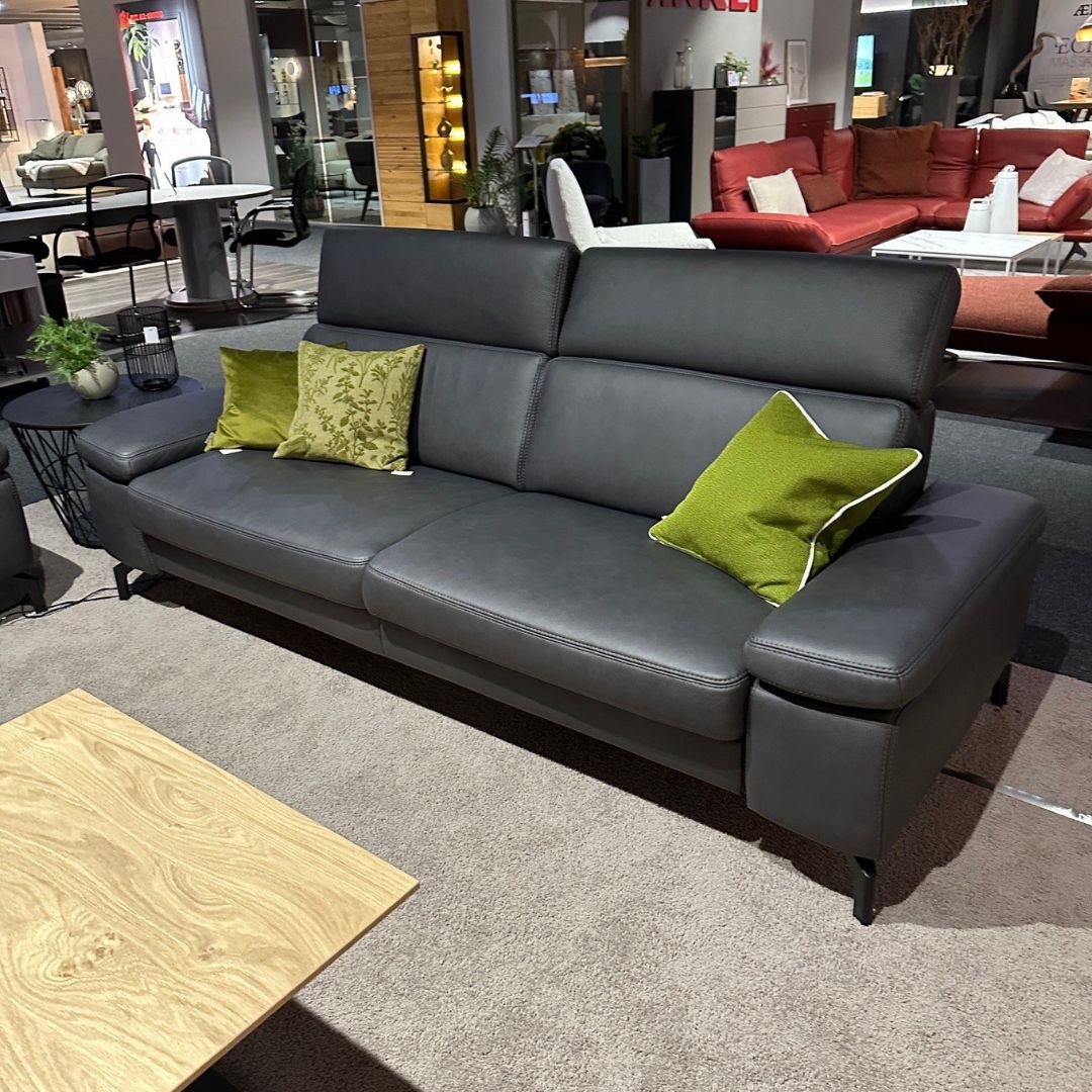 Ein modernes graues Ledersofa mit grünen Kissen, umgeben von einer stilvollen Wohnzimmerumgebung im Möbelgeschäft.
