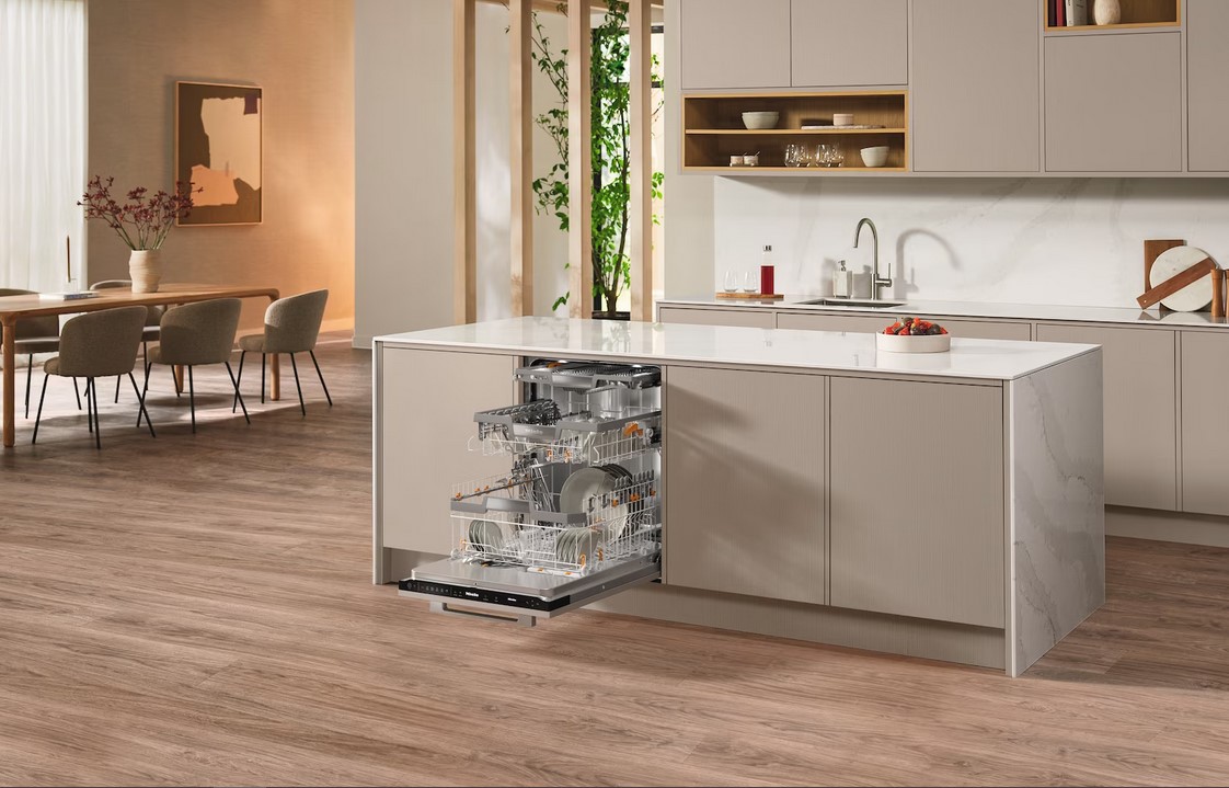 Eine offene, vollintegrierte Miele Spülmaschine in einer modernen, hellen Küche. Die Körbe der Spülmaschine sind mit Geschirr gefüllt und herausgezogen. Im Hintergrund ist ein Essbereich mit einem Holztisch und Stühlen zu sehen.