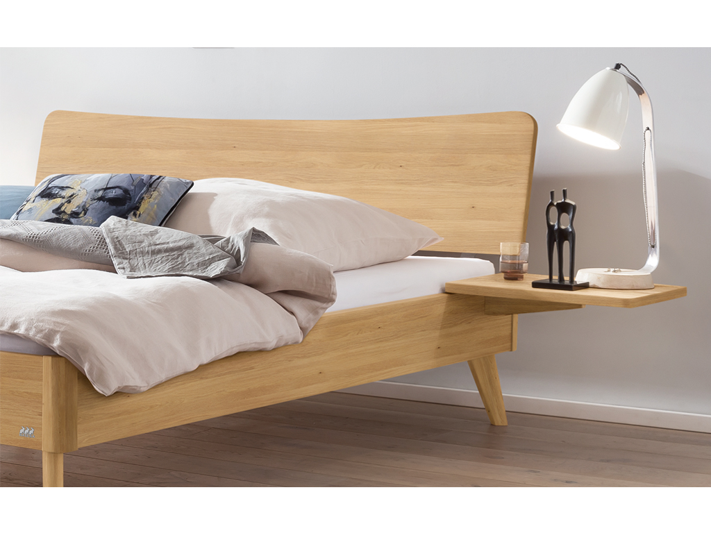 Ein Hasena Bett Caluso aus hellem Eichenholz ("Eiche bright"). Das Bett hat ein geschwungenes Kopfteil und schräg ausgestellte Beine. An der Seite ist ein schwebender Nachttisch mit einer Lampe und einer kleinen Skulptur zu sehen.
