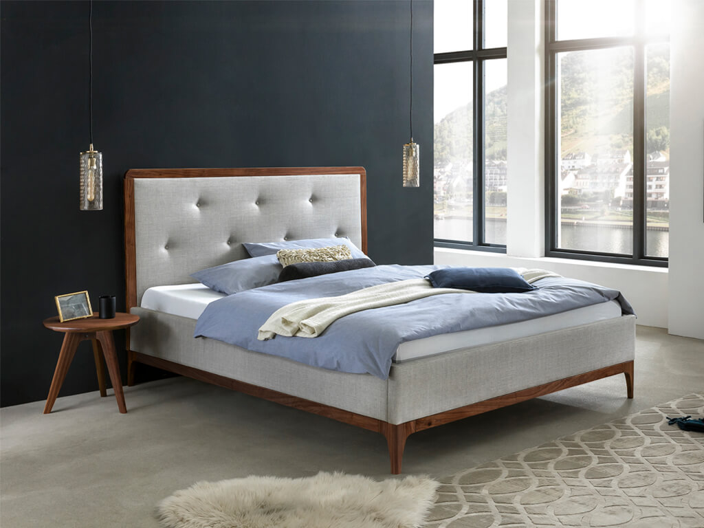 Ein Bett mit hellem, grauem Stoffbezug und einem Holzrahmen in Walnuss, das in einem modern eingerichteten Schlafzimmer mit großen Fenstern steht.