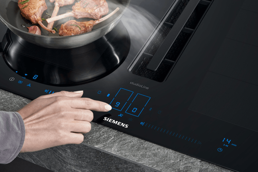 Nahaufnahme eines modernen Induktionsherds mit Touch-Bedienung. Eine Pfanne mit brennenden Lammkoteletts steht auf dem Herd.