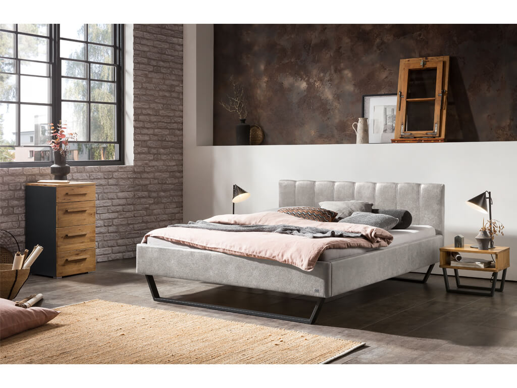 Ein modernes Schlafzimmer mit einem grauen Polsterbett, passendem Nachttisch und einer Kommode vor einer Backsteinwand.