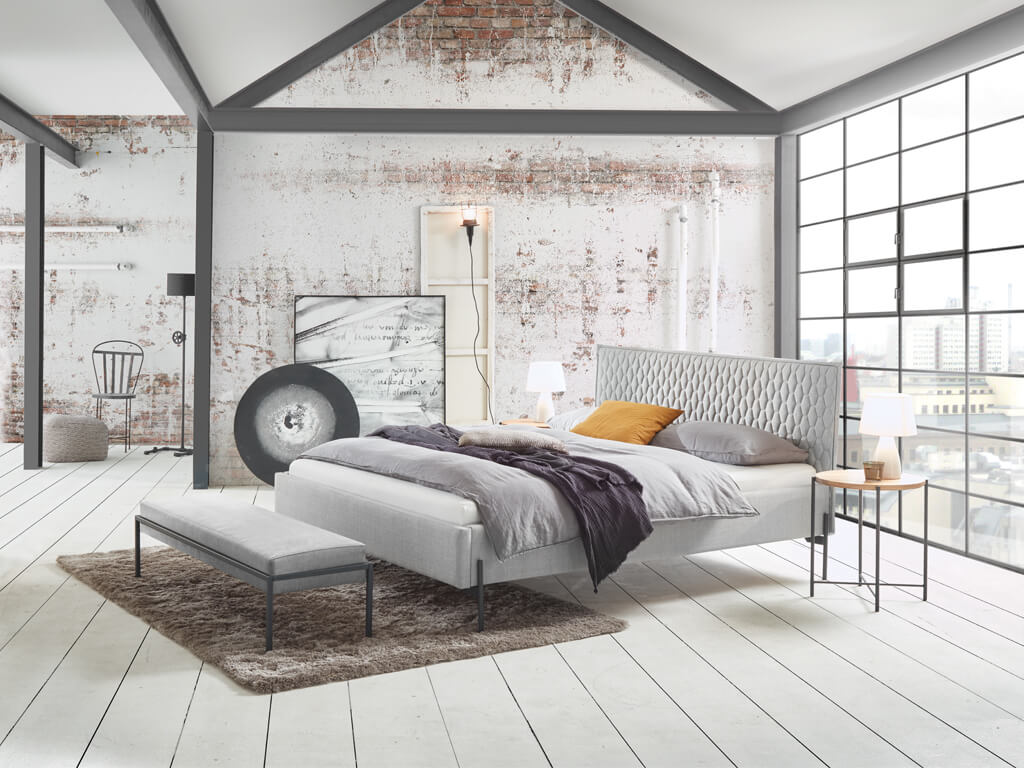 Ein glamouröses Schlafzimmer-Set, das das Bett Gloria mit einem gesteppten Kopfteil, einer passenden Bank und einem Hocker in einem modernen Loft-Stil zeigt.