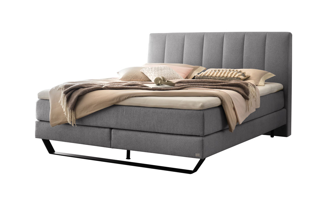 graues Boxspringbett mit Stahlkufe in schwarz, stilvoll dekoriert mit Decken und Kissen vor weißem Hintergrund