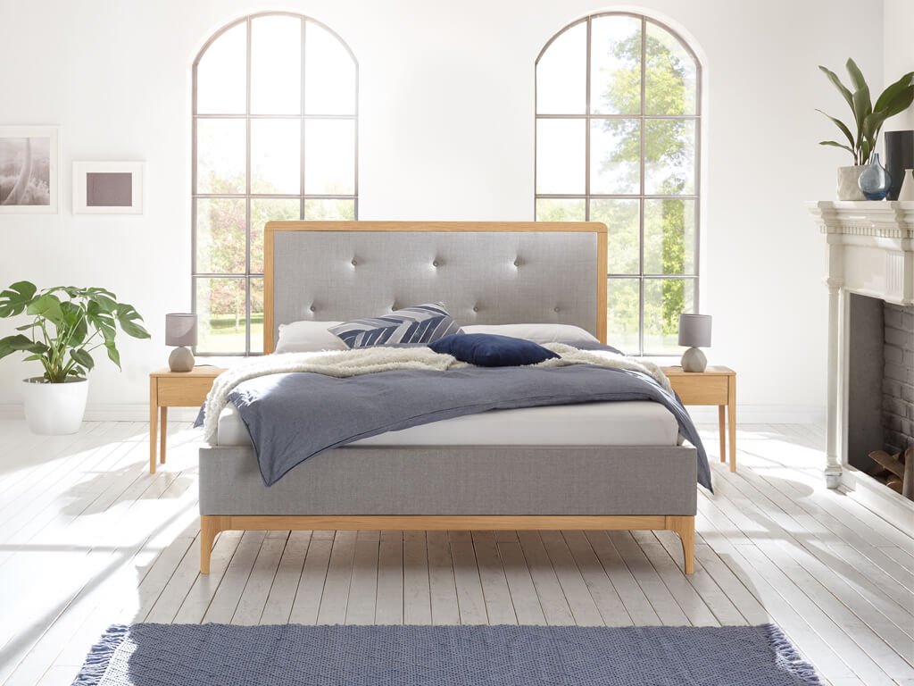 Ein Bett mit hellem Holzrahmen und grauem Stoffbezug in einem hellen, sonnendurchfluteten Schlafzimmer mit zwei großen Bogenfenstern.