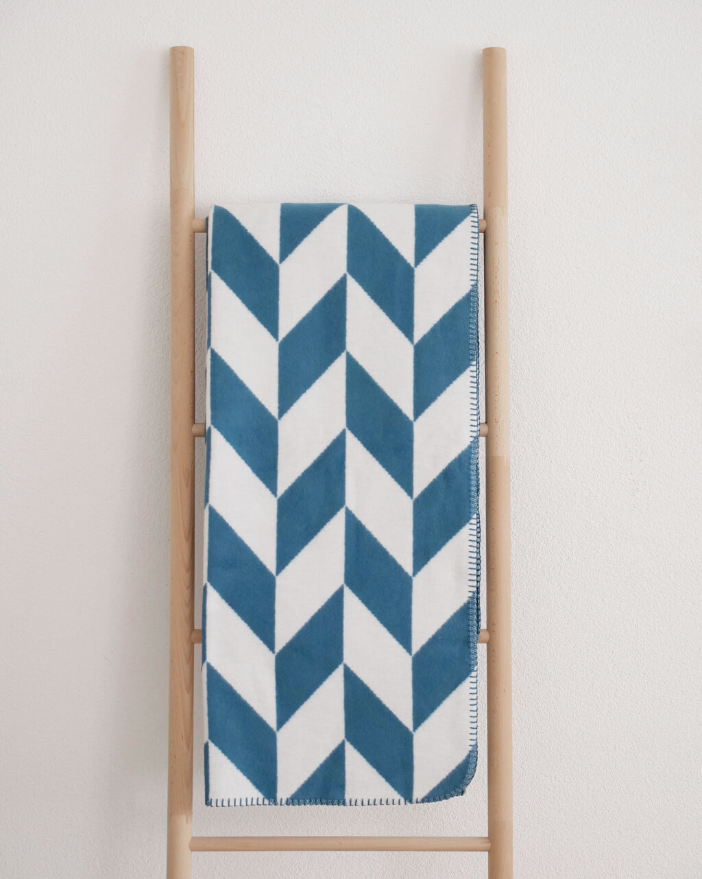 RIEMA MADS – Wohndecke, Bio-Baumwolle - Pfeile | 150 x 200 cm | blau-weiß Blau-weiße Decke mit Zickzack-Muster, elegant präsentiert auf einfacher Holzleiter an einer weißen Wand