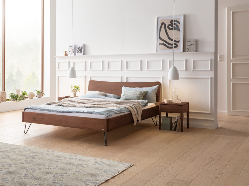 Ein Hasena Bett Locarno aus Walnuss-Holz (Nussbaum), in einem hellen Schlafzimmer mit minimalistischer Dekoration und Haarnadel-Beinen in Anthrazit. Auf der rechten Seite steht ein passender Nachttisch.