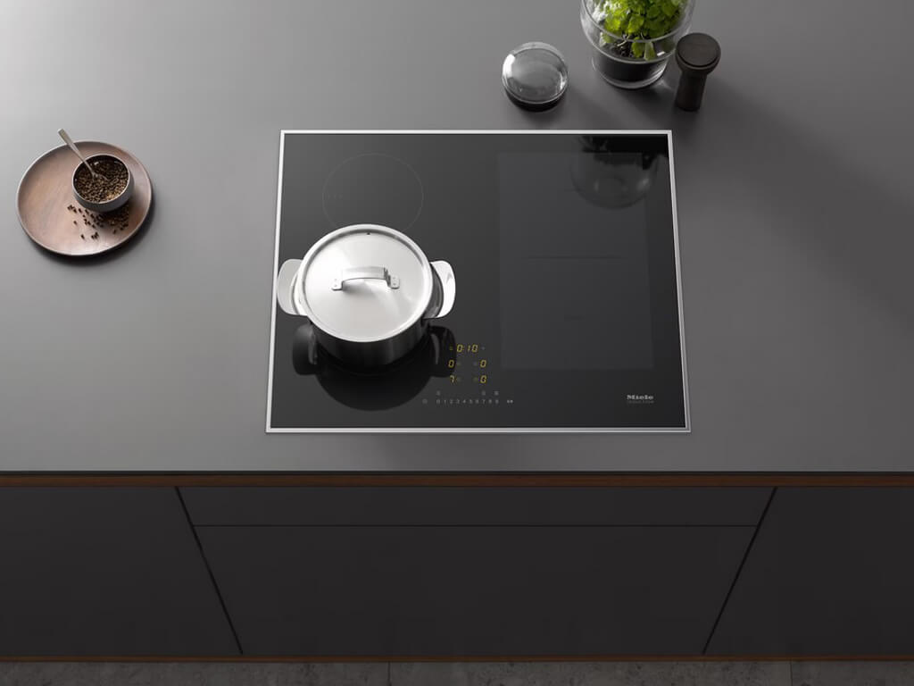 Draufsicht auf ein Miele Induktionskochfeld, das in eine graue Arbeitsplatte eingelassen ist. Ein silberner Kochtopf steht auf einer der runden Kochzonen auf der linken Seite.