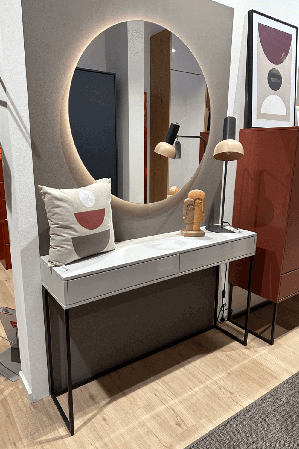 Ein stilvoller Schminktisch mit rundem, beleuchtetem Spiegel, minimalistischer Tischlampe, modernem Kissen und einer Skulptur, platziert auf hellem Holzboden in einem modernen Raum.