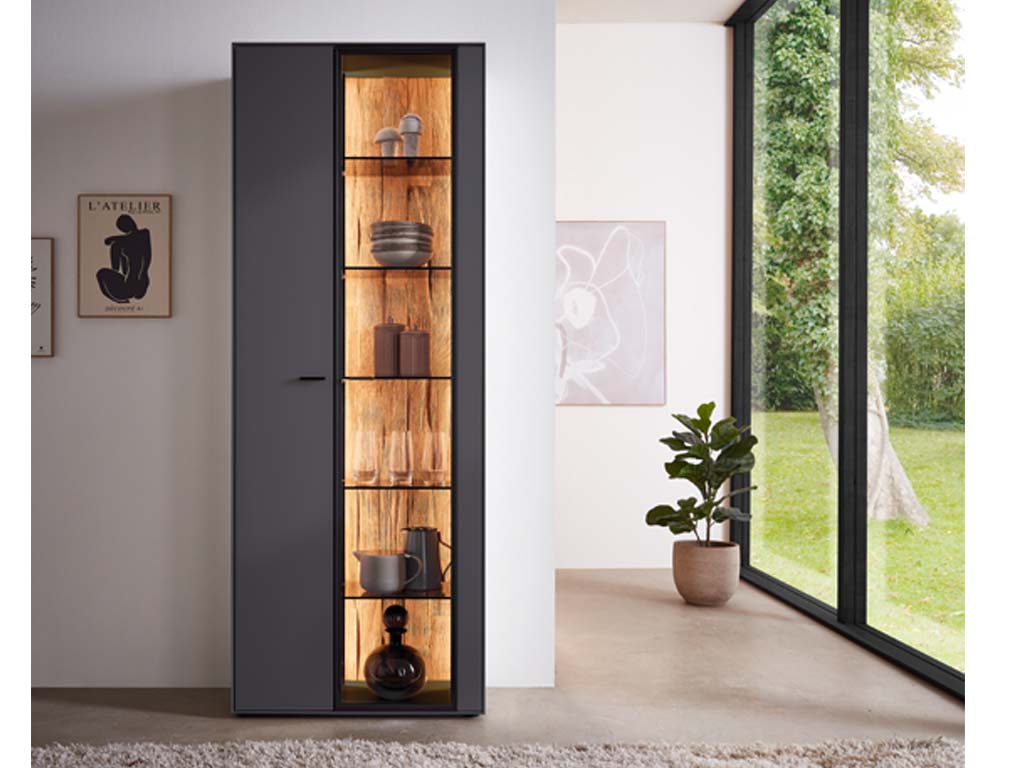Wöstmann WM 2470 - Vitrine | Type 6743/6744 | B: 76,9 cm Ein eleganter schwarzer Schrank mit Glasfront, beleuchtet, im modernen Wohnzimmer neben einem großen Fenster mit Pflanzen und dekorativen Elementen auf den Regalen.