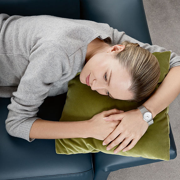 Eine Frau liegt seitlich auf einem Sofa, den Kopf auf ein grünes Kissen gebettet und trägt einen grauen Pullover sowie eine schlichte Armbanduhr.