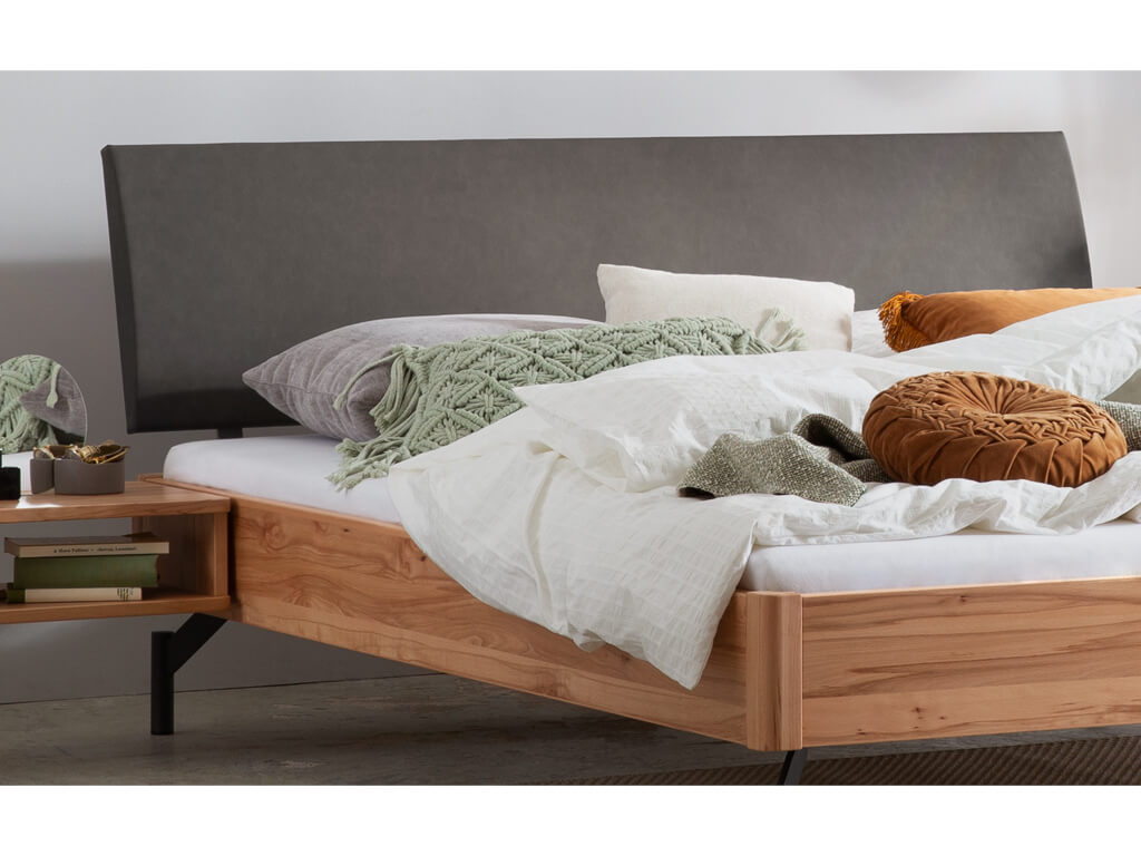 Ein Detailbild des Bettes "Broni" mit einem Rahmen aus Wildbuchenholz. Das Bett hat ein gepolstertes anthrazitfarbenes Kopfteil und dunkle Metallbeine. Ein passender Nachttisch aus Wildbuchenholz steht ebenfalls neben dem Bett.