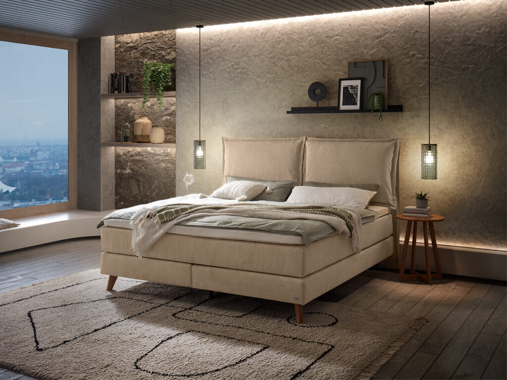 Ein Hasena Boxspringbett Pronto Ricarda Roy mit einem sandfarbenen Stoffbezug in einem gemütlich beleuchteten Schlafzimmer mit Holzfußboden.