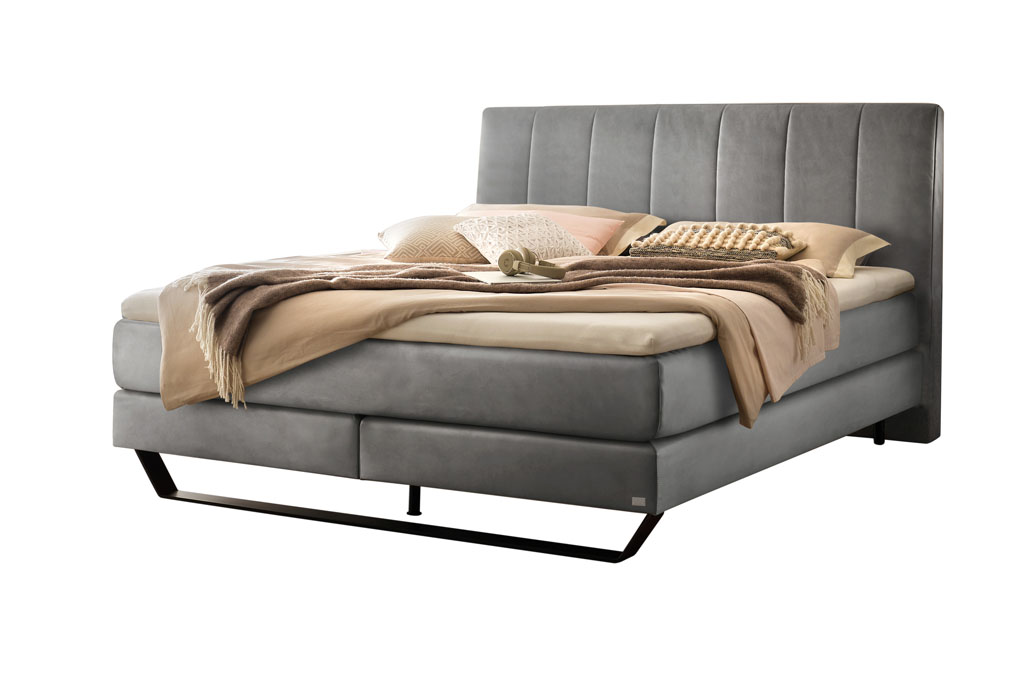graues Boxspringbett mit Stahlkufe in schwarz, stilvoll dekoriert mit Decken und Kissen vor weißem Hintergrund