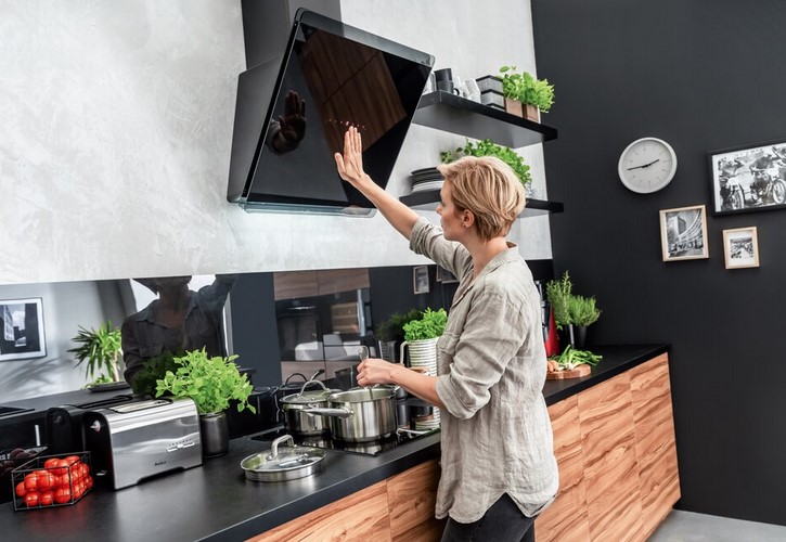 Eine Frau steht in einer modernen Küche und bedient die Touch-Steuerung einer schwarzen, schrägen Amica Dunstabzugshaube über einem Kochfeld.