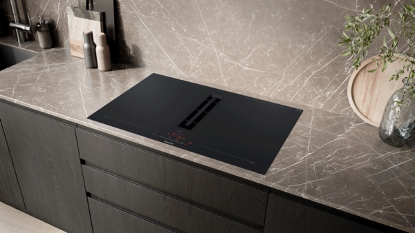 Das schwarze Siemens StudioLine Induktionskochfeld mit integriertem Dunstabzug ist in eine moderne Küchenarbeitsplatte aus braun-grau meliertem Naturstein oder Marmor eingelassen. Die Umgebung zeigt dunkle Holzschubladen und eine passende Rückwand.