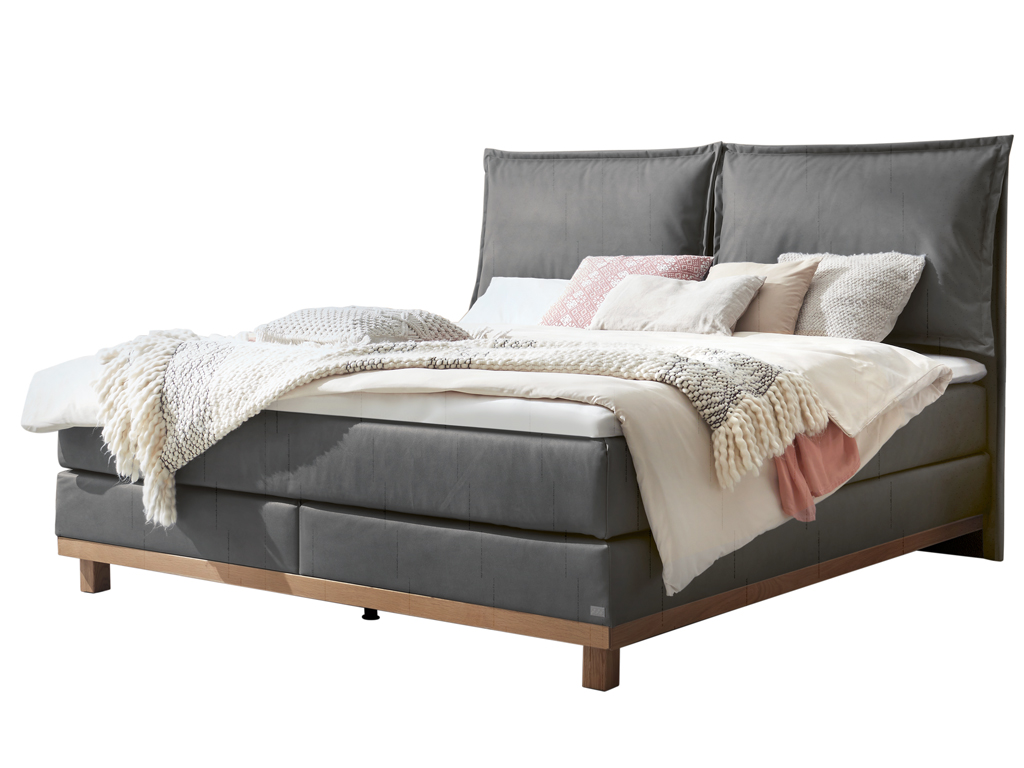 Ein freigestelltes Produktfoto des Boxspringbettes Hasena Pronto Zamora mit stahlgrauem Stoffbezug, dekoriert mit Kissen und einer Decke.