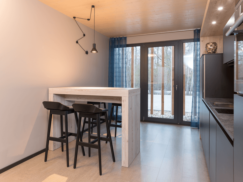 Minimalistisch eingerichtete Küche mit weißem Holztisch, schwarzen Barhockern und großen Fenstern mit blauen Vorhängen