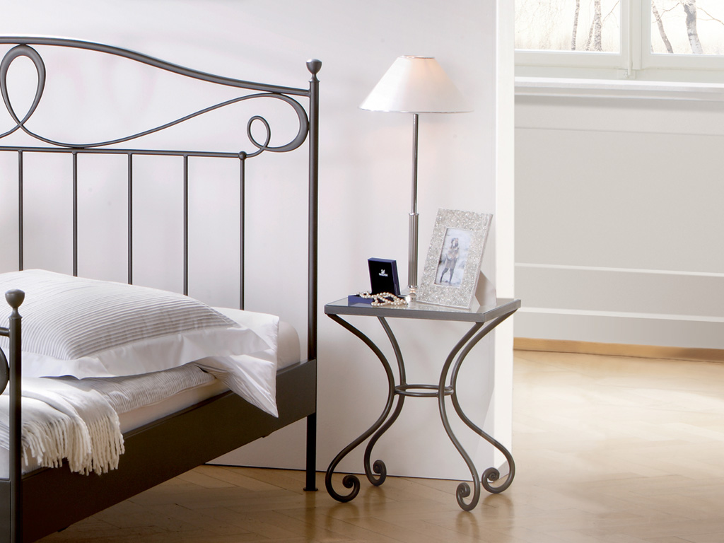 schwarzer Nachttisch auf filigranem Eisenmetallgestell mit Glasplatte an einem Metallbett mit weißer Bettwäsche in einem stilvollen Zimmer Cura von Hasena