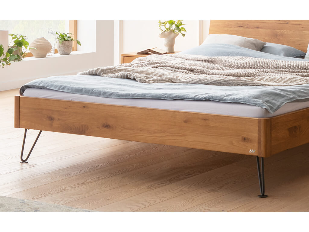 Nahaufnahme des unteren Teils eines Hasena Bett Locarno mit einem Rahmen in Wildeiche natur geölt und schwarzen, anthrazitfarbenen Haarnadel-Beinen. Man sieht den Bettrahmen, die Maserung des Holzes, die Beine und den Holzboden.