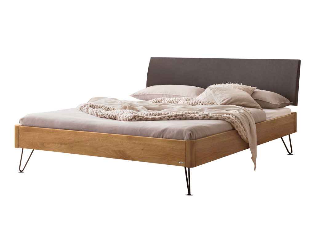 Freigestelltes Bett mit einem Bettrahmen aus naturbelassener geölter Wildeiche, einem anthrazitfarbenen Kopfteil und schwarzen, schmalen Haarnadelbeinen. Das Bett ist mit heller Bettwäsche und einem groben Strickplaid dekoriert.