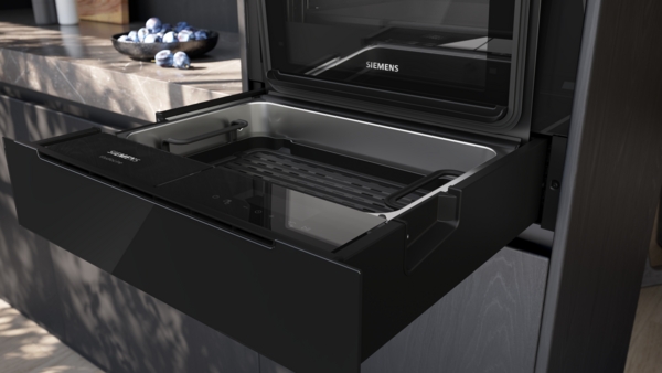 Nahaufnahme einer offenen, ausziehbaren, schwarzen Siemens StudioLine Schublade. Im Inneren ist eine silberne Wanne mit einem schwarzen Grillrost und einer dunklen Steuerungsleiste zu sehen.