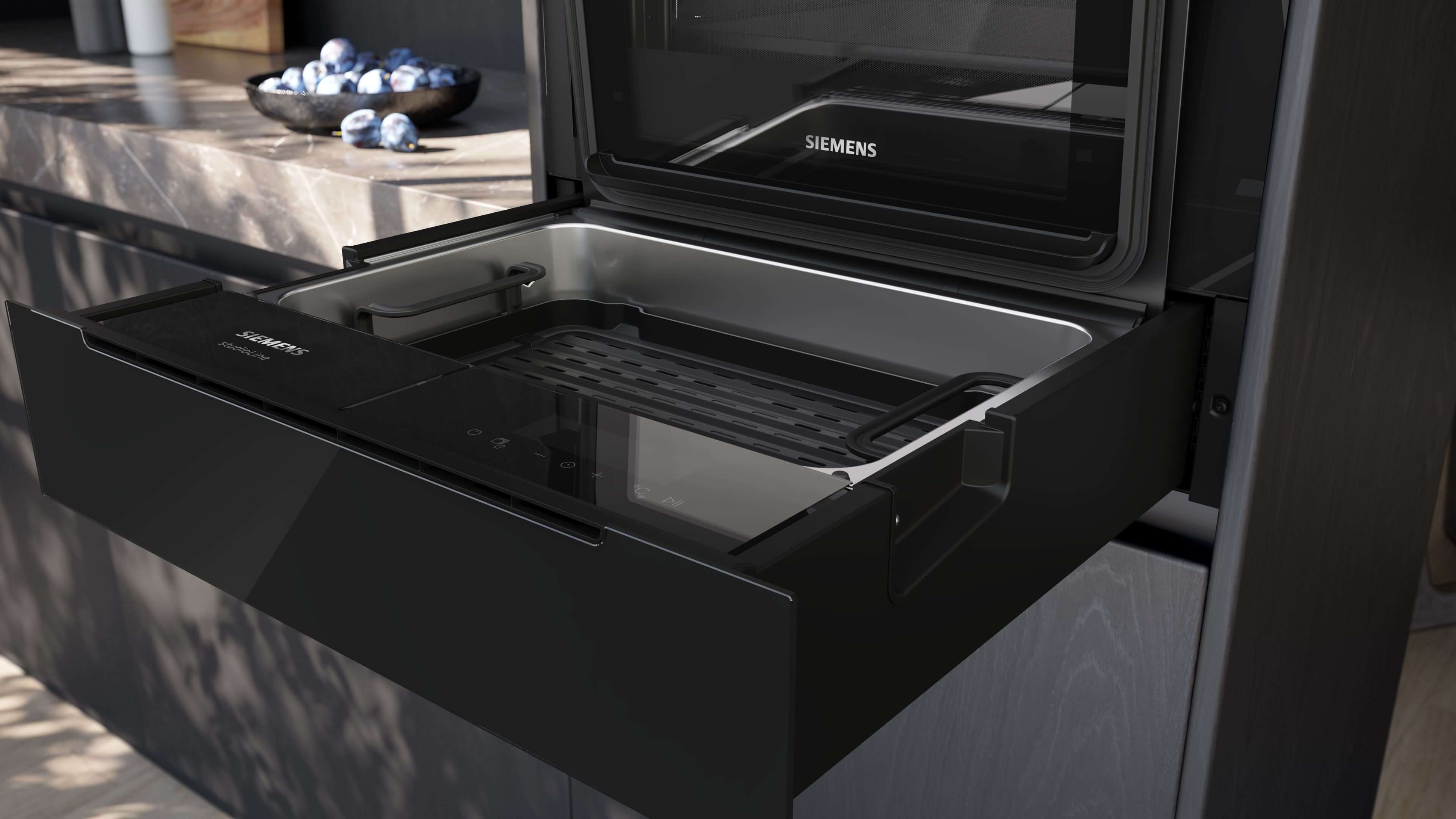 Nahaufnahme einer offenen, ausziehbaren, schwarzen Siemens StudioLine Schublade. Im Inneren ist eine silberne Wanne mit einem schwarzen Grillrost und einer dunklen Steuerungsleiste zu sehen.