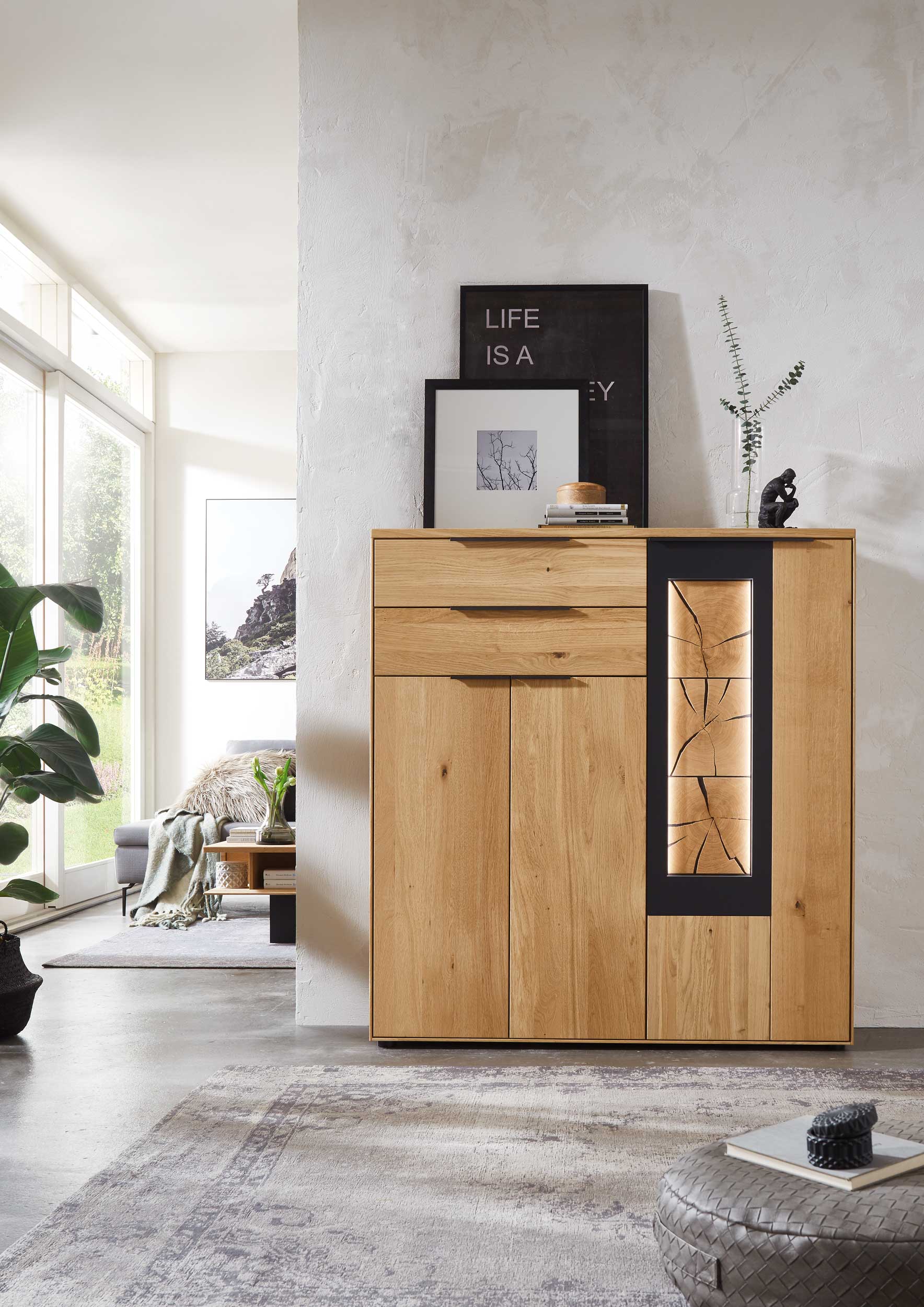 Wöstmann WM 1910 - Highboard | Type 4253/4254 | Wildeiche massiv | B: 123,9 cm Ein holzfarbener Sideboard-Schrank steht in einem modernen Wohnzimmer mit heller Einrichtung und Dekoration.