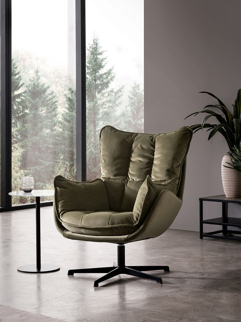 designwerk Jules - Sessel Eleganter grüner Samtsessel vor einem Fenster mit Blick auf Bäume, neben einem kleinen runden Beistelltisch
